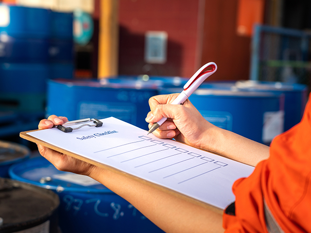 Hazard Analysis Safety inspection worker holding a pen and clipboard with a checklist document attached, a blurred background of chemical storage area