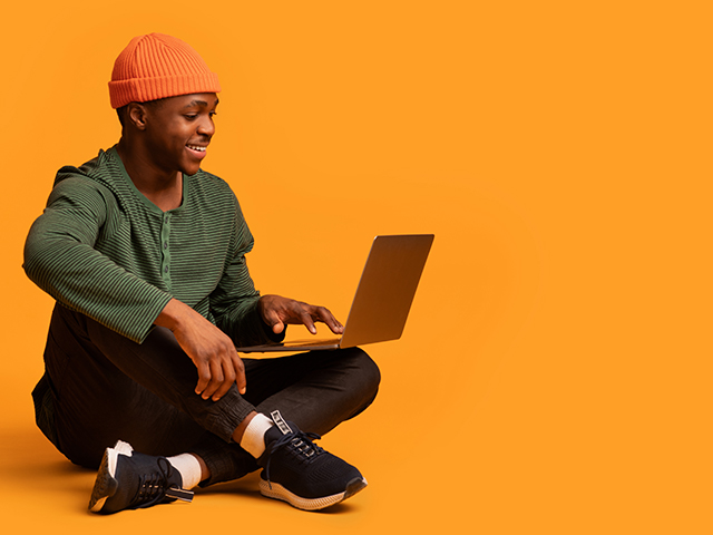 A beanie-clad African American male student sits on the floor, cross-legged, using a laptop, with a cheerful expression against an orange backdrop.