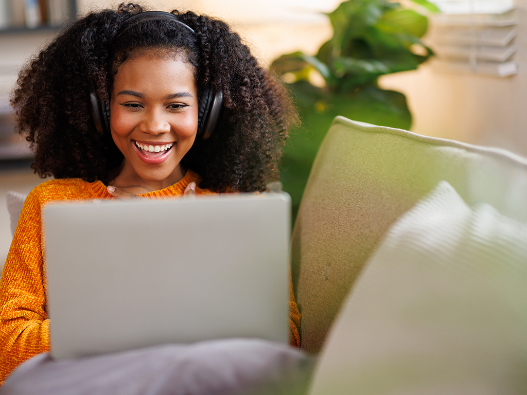 African American young woman student sits on couch, using a laptop, with a cheerful expression