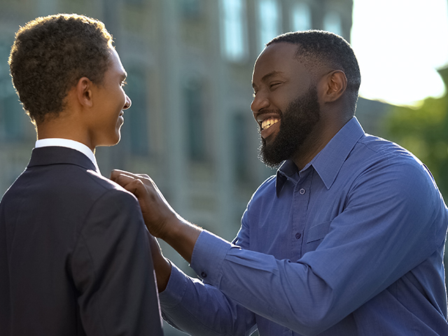 smiling father/ guardian helping young adult son with tie