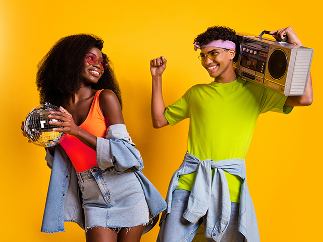 Photo of two young adults dressed denim outfits dancing holding boom box disco ball in front of an orange color background