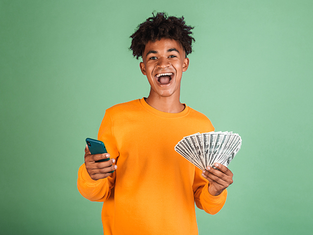 smiling young man holding big fan of money in one hand and a smart phone in the other hand, in front of green color background